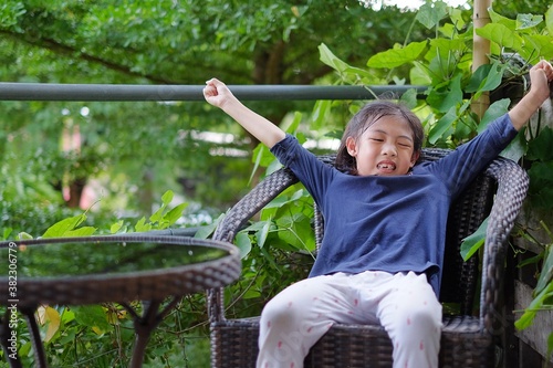 A cute young Asian girl sitting on a big ratton chair feeling bored and sleepy, stretching her arms upward with her eyes closed. photo