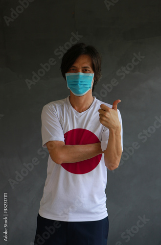 Man wearing hygienic mask and wearing Jananese flag colored shirt and cross one s arm with thumbs up on dark wall background. photo