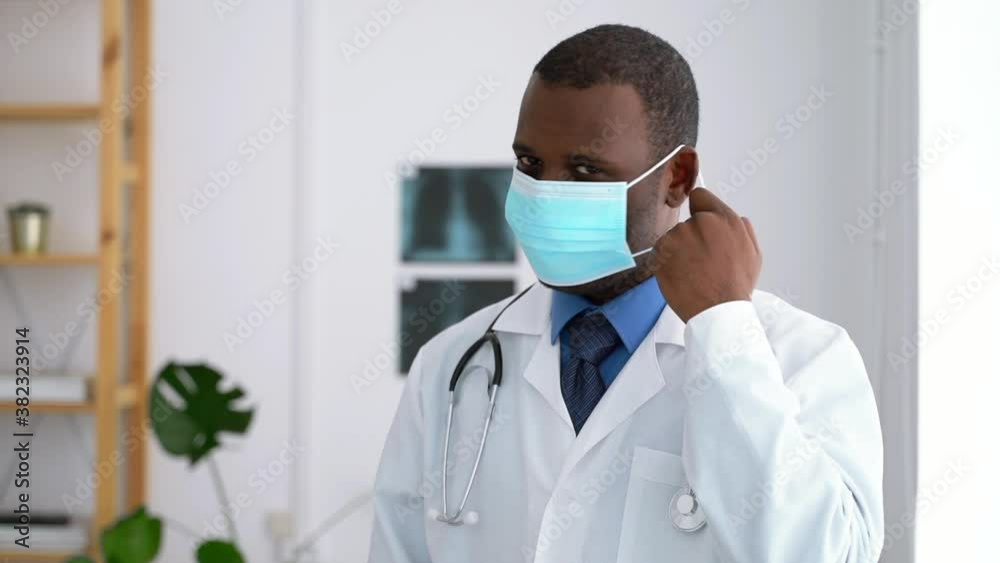 Doctor mask portrait young male takes off protective mask and poses while working at modern clinic spbas. Front view of one African American man posing for camera with smile, standing in bright room