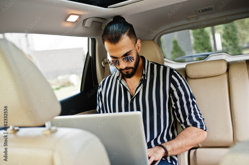 Successful arab man wear in striped shirt and sunglasses pose inside white suv car with laptop in hands.