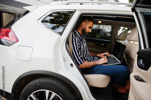 Successful arab man wear in striped shirt and sunglasses pose inside white suv car with laptop in hands. © AS Photo Family
