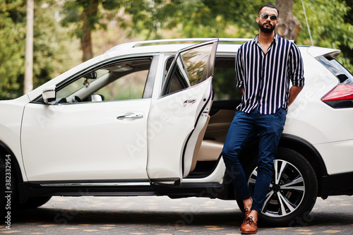 Successful arab man wear in striped shirt and sunglasses pose near his white suv car. Stylish arabian men in transport. © AS Photo Family