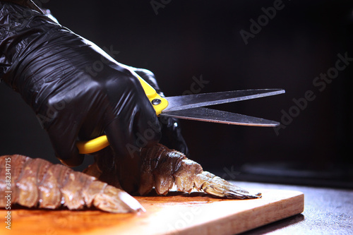 Cooking a dish of langoustes. The chef in black gloves with a pair of scissors cut up the lobster tails. Unrecognizable photo. Only hand. Black background. Close up.