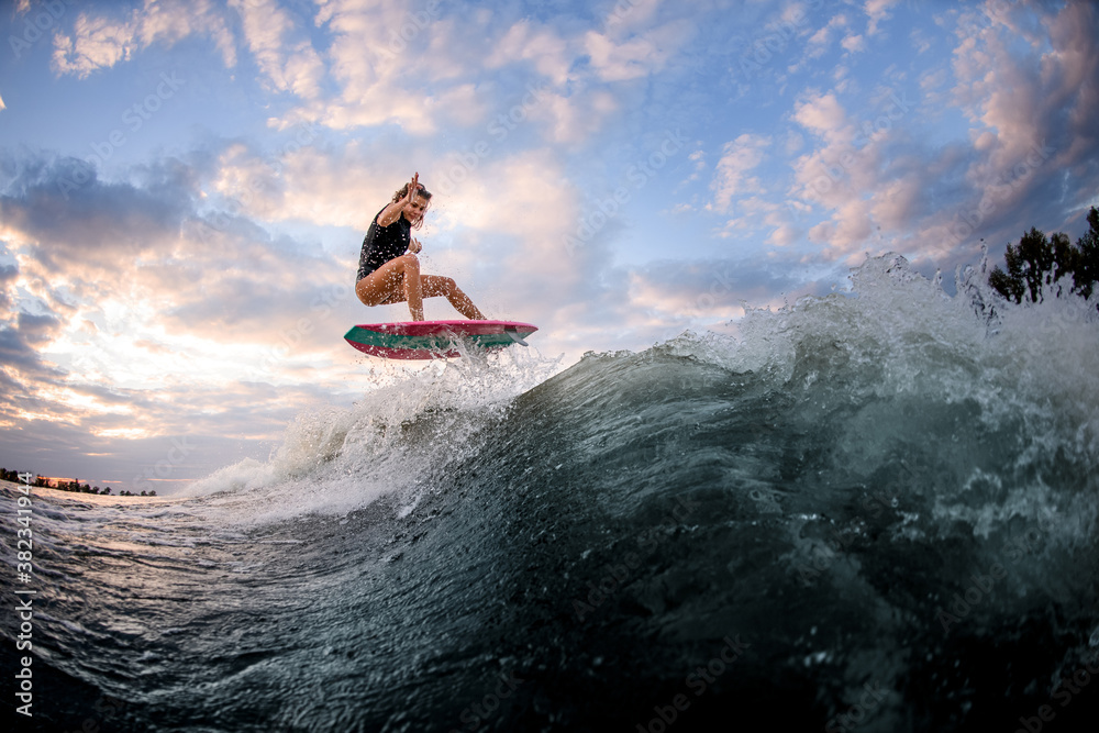 athletic young woman in swimsuit jumping up on surf style wakeboard against blue cloudy sky