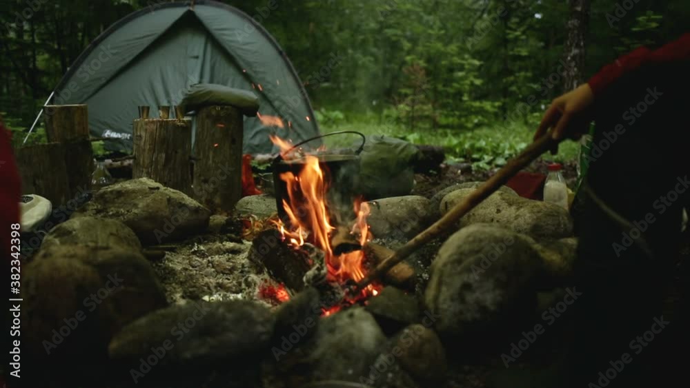 Young man is making bonfire while sitting at camping on summer day iroi. Closeup view of male tourist stirs coals of campfire with wooden stick, sits by stones in cloudy weather. One traveler has good Young man is making bonfire while sitting at camping on summer day iroi. Closeup view of male tourist stirs coals of campfire with wooden stick, sits by stones in cloudy weather. One traveler has good