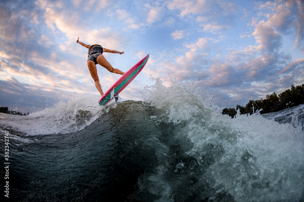 Rear view of young attractive woman who stands on surf style wakeboard and jumps on wave.