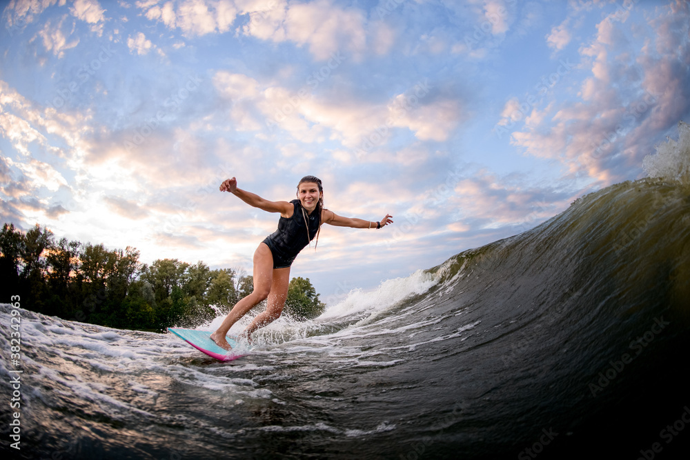 smiling young woman stands on bright surf style wakeboard and rides down big water wave