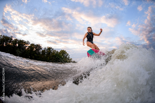 cheerful young woman stands on bright surf style wakeboard and rides big splashing wave © fesenko