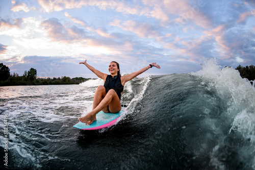 cheerful woman riding wave while sitting on surf style wakeboard with outstretched arms