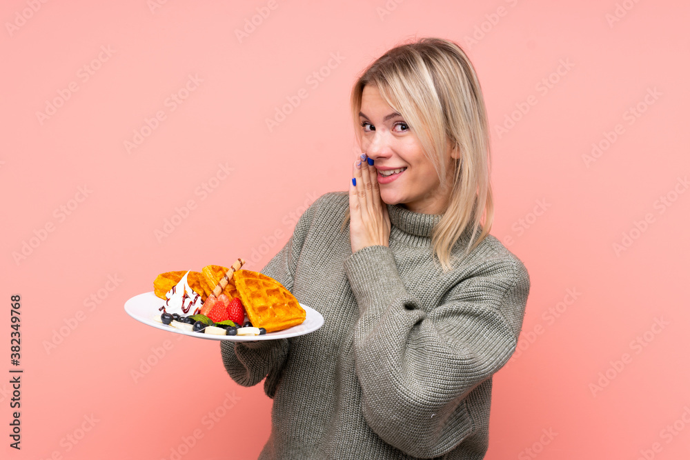 Young blonde woman holding waffles over isolated pink background whispering something