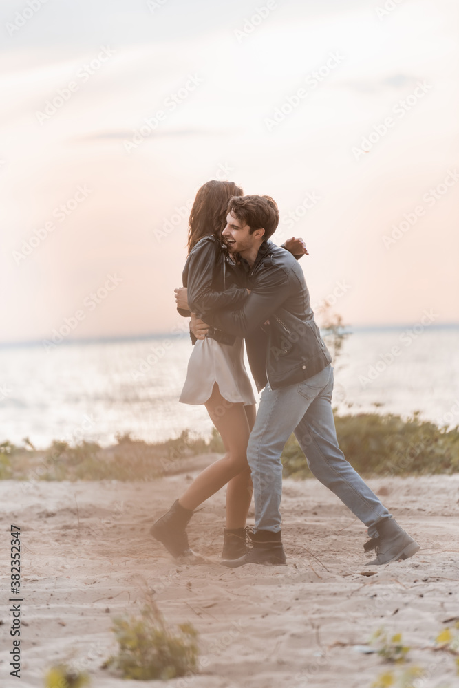 Selective focus of man in leather jacket hugging girlfriend in dress on beach at sunset