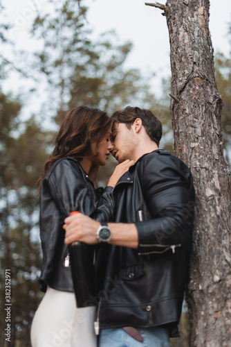 Selective focus of woman kissing boyfriend with bottle of wine near tree in forest