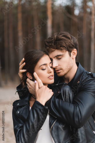 Young man with closed eyes embracing brunette girlfriend in leather jacket near forest