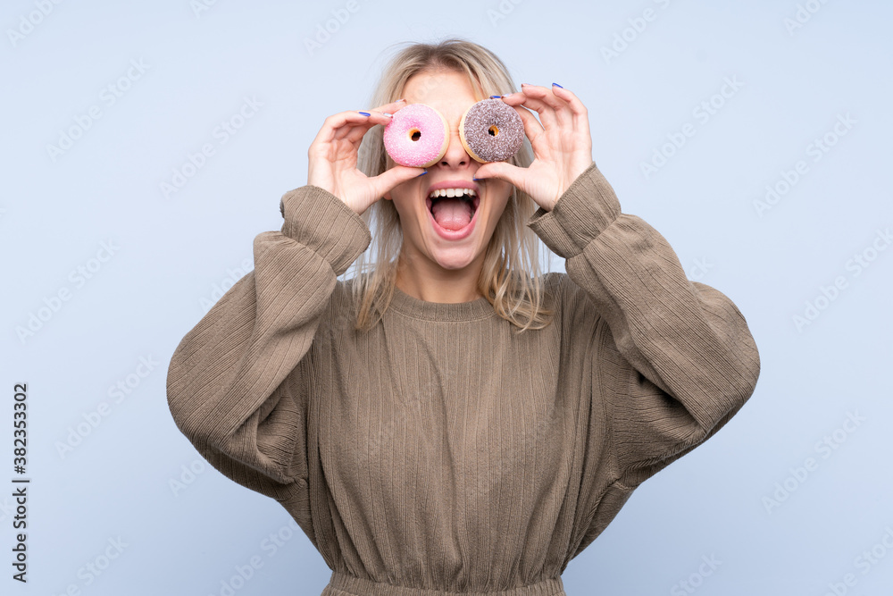Young blonde woman over isolated blue background holding donuts in an eye