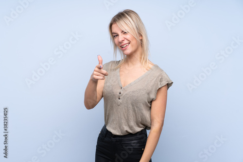 Young blonde woman over isolated blue background points finger at you