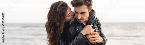 Panoramic shot of young woman embracing boyfriend in leather jacket beside sea