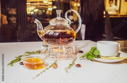 Aromatic hot green tea in a glass teapot with a warm blurry background with honey and fresh mint leaves. Served table with tea in a cafe. Kettle warmer