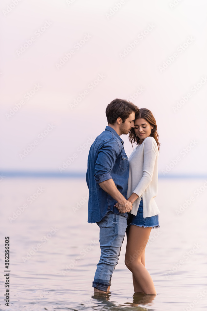 Side view of young couple holding hands while standing in sea at evening Side view of young couple holding hands while standing in sea at evening