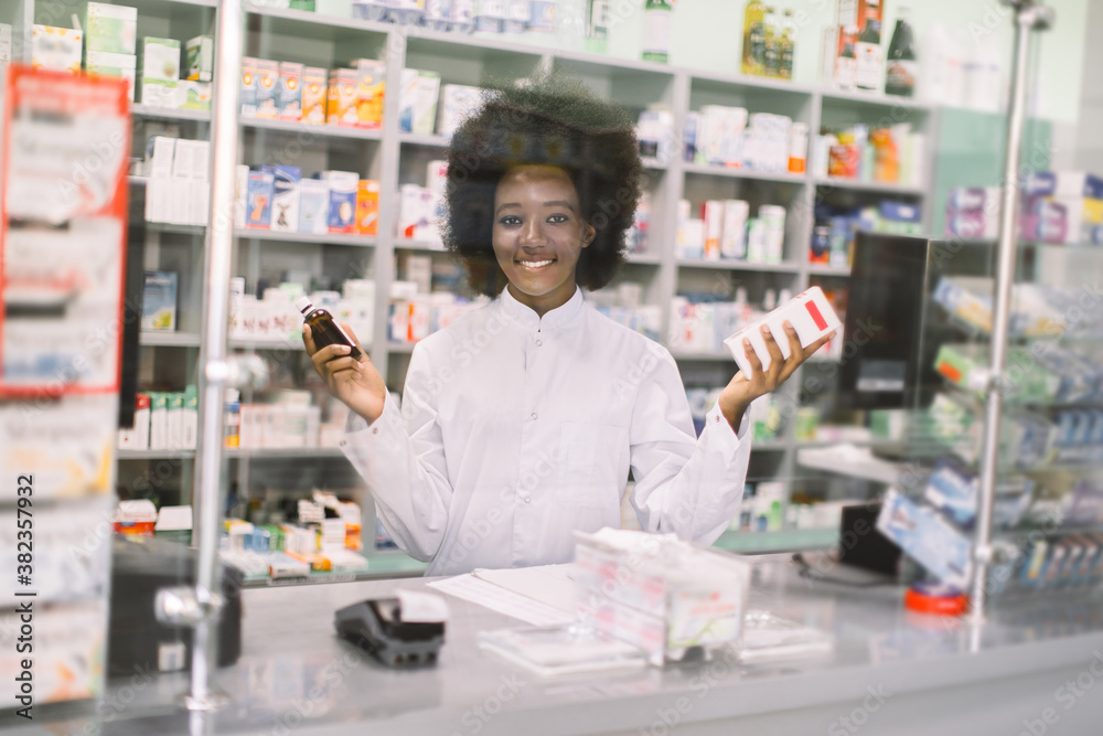 Portrait of pretty young African pharmacist behind the glass, smiling at camera in modern drugstore at the counter, holding medications in her hands. Pharmacy and medicines concept