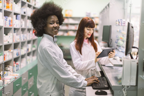 Two young cheerful pharmacists women working together at pharmacy. African woman typing on computer while her Caucasian colleague using tablet. Focus on smiling African woman
