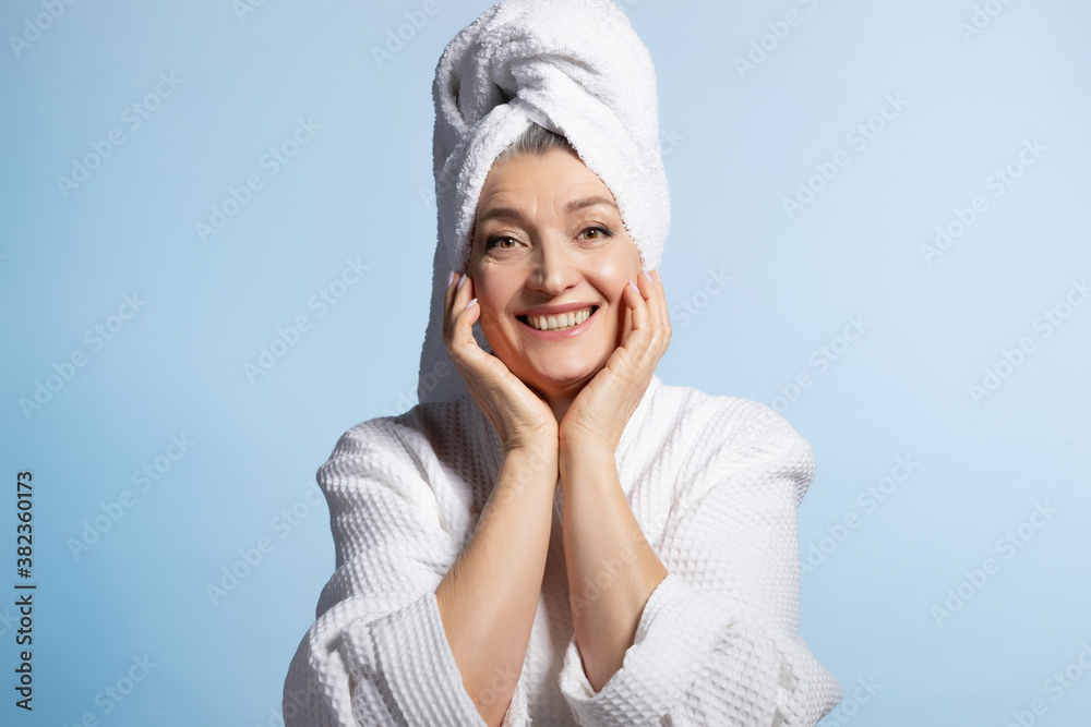 Pleased elderly woman in towel and housecoat touching her cheeks and smiling with tooth on blue studio background. Anti aging skincare concept. Portrait of friendly senior female after taking shower. Pleased elderly woman in towel and housecoat touching her cheeks and smiling with tooth on blue studio background. Anti aging skincare concept. Portrait of friendly senior female after taking shower.