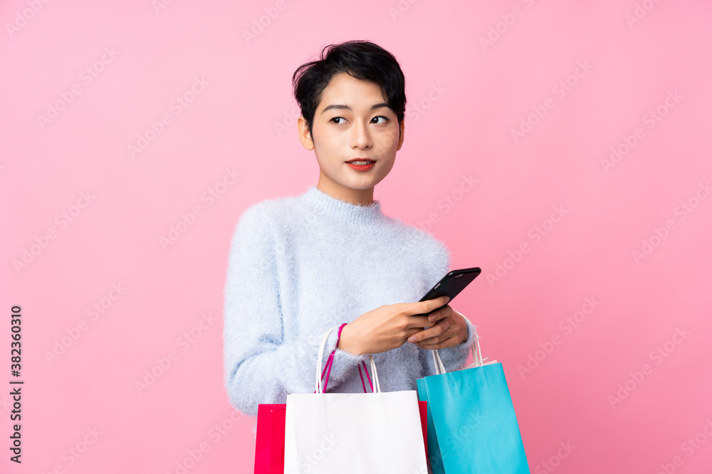 Young Asian girl over isolated pink background holding shopping bags and writing a message with her cell phone to a friend Young Asian girl over isolated pink background holding shopping bags and writing a message with her cell phone to a friend