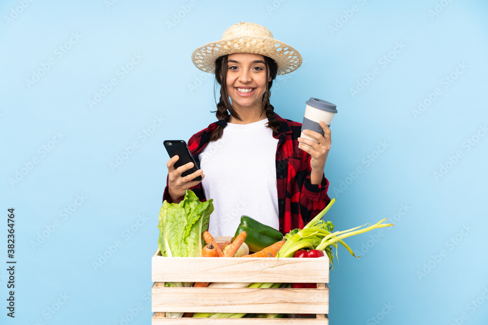 Young farmer Woman holding fresh vegetables in a wooden basket holding coffee to take away and a mobile