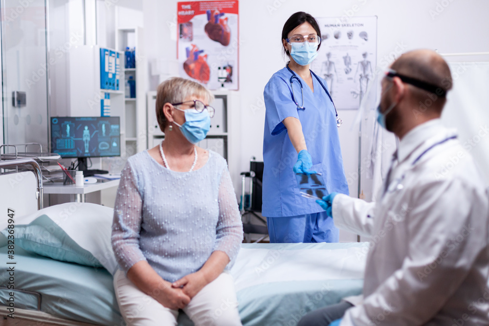 Fototapeta premium Nurse giving doctor x-ray of senior woman patient in hospital examination room wearing face mask against coronavirus outbreak. Medical examination for infections, disease and diagnosis.
