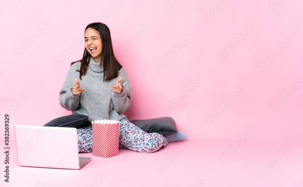 Young mixed race woman eating popcorn while watching a movie on the laptop pointing to the front and smiling Young mixed race woman eating popcorn while watching a movie on the laptop pointing to the front and smiling