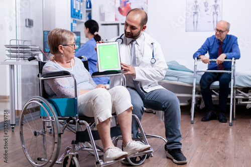 Medical practition holding tablet pc with green screen during consultation of disabled senior woman in wheelchair. Man with disabilities ,walking frame sitting in hospital bed. Health care system photo