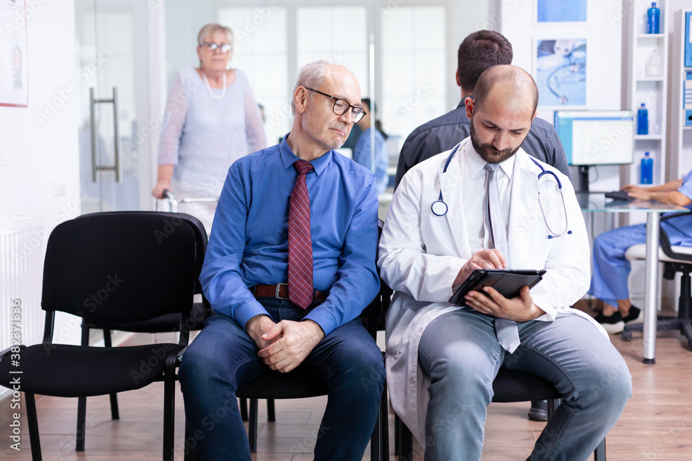 Doctor explaining test results to senior man in hospital waiting area. Disabled senior woman with walking frame arriving for medical examination. Doctor explaining test results to senior man in hospital waiting area. Disabled senior woman with walking frame arriving for medical examination.