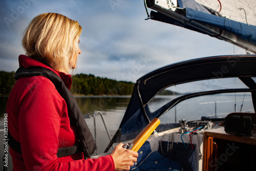 Blonde senior woman steering her sailboat in the summer sunset out on the sea wearing a life jacket
