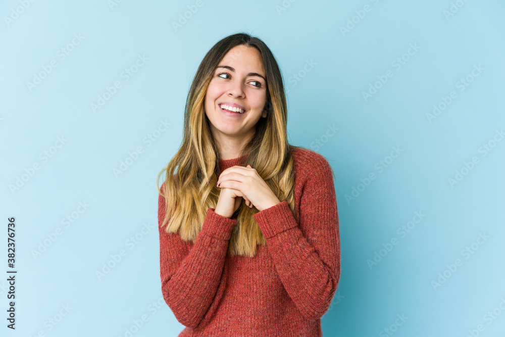 Young caucasian woman isolated on blue background keeps hands under chin, is looking happily aside. Young caucasian woman isolated on blue background keeps hands under chin, is looking happily aside.