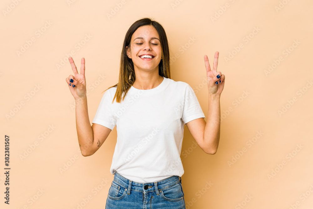 Young woman isolated on beige background showing victory sign and smiling broadly. Young woman isolated on beige background showing victory sign and smiling broadly.