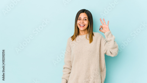 Young woman isolated on blue background winks an eye and holds an okay gesture with hand.