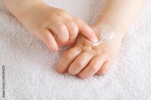 Baby girl finger applying moisturizing cream on hand on white towel. Care about children clean and soft body skin. Front view. Closeup.