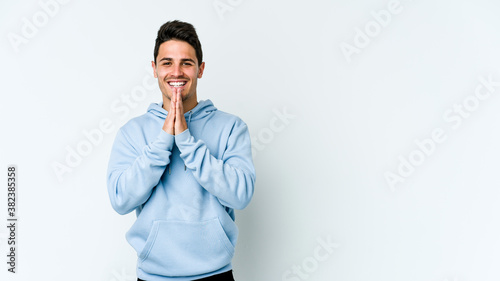 Young caucasian man isolated on white background holding hands in pray near mouth, feels confident.