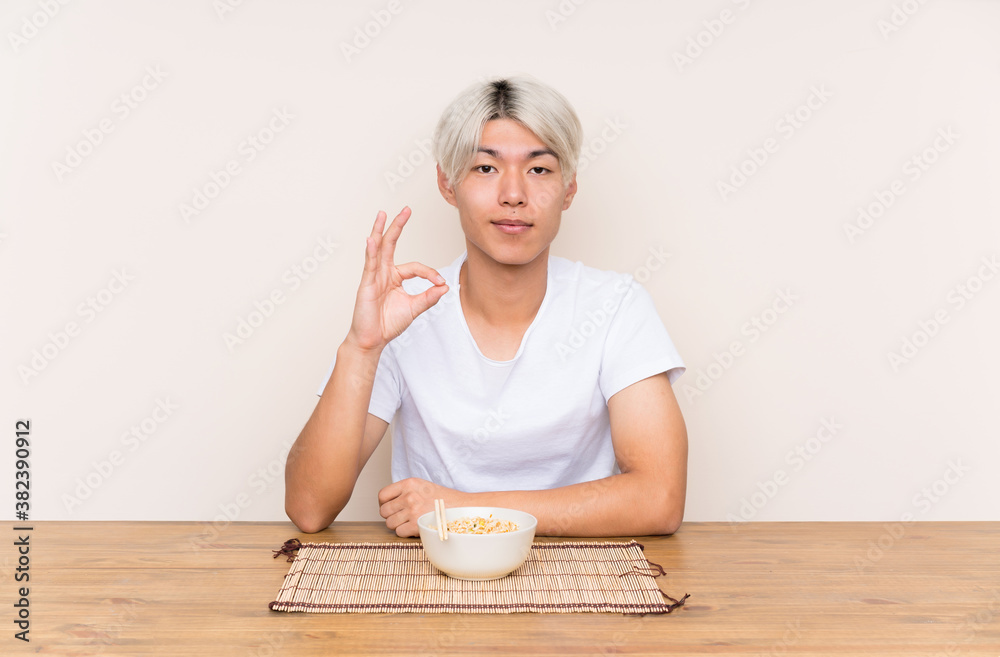 Young asian man with ramen in a table showing an ok sign with fingers Young asian man with ramen in a table showing an ok sign with fingers