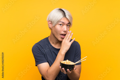 Young asian man over isolated yellow background whispering something