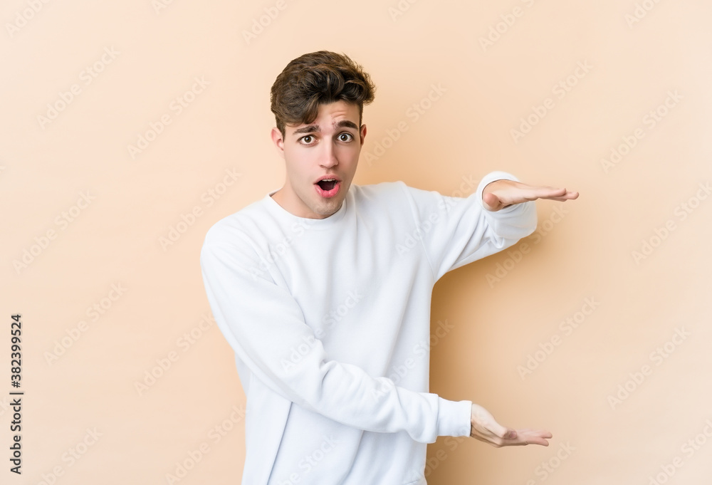 Young caucasian man isolated on beige background shocked and amazed holding a copy space between hands. Young caucasian man isolated on beige background shocked and amazed holding a copy space between hands.