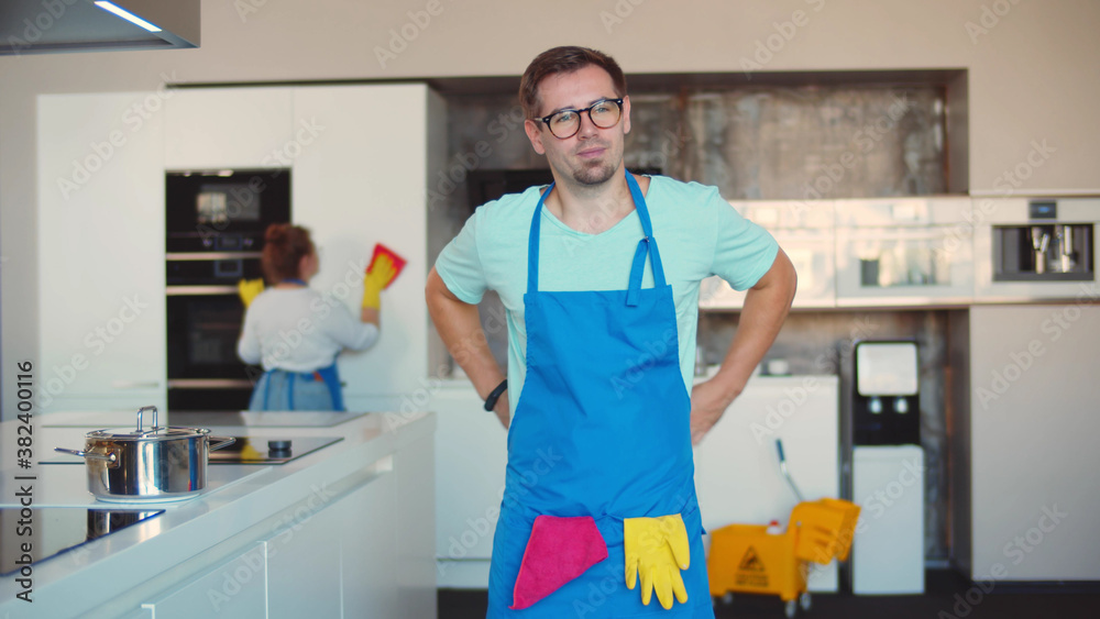 Man janitor putting on apron preparing for work in kitchen with colleague cleaning on background Man janitor putting on apron preparing for work in kitchen with colleague cleaning on background