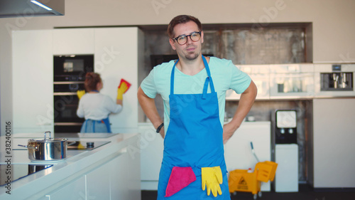 Man janitor putting on apron preparing for work in kitchen with colleague cleaning on background