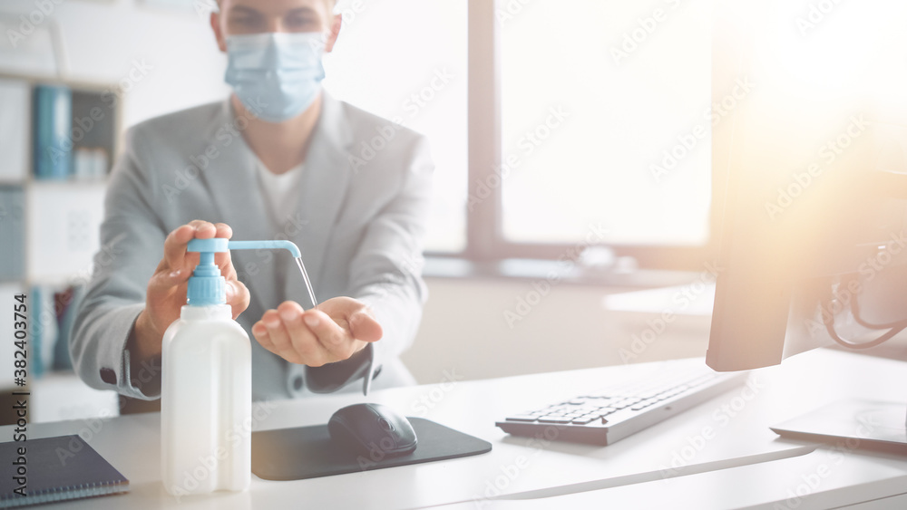 Handsome Stylish Young Man Wearing Protective Face Mask Uses Hand Sanitizer while Working at His Desk on a Computer in Office. Blue Eyed Blond Haired Young Male Follows Health and Safety Instructions Handsome Stylish Young Man Wearing Protective Face Mask Uses Hand Sanitizer while Working at His Desk on a Computer in Office. Blue Eyed Blond Haired Young Male Follows Health and Safety Instructions