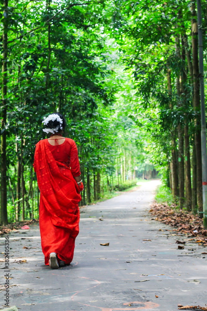 Young woman wearing red saree walking on green asphalt road. The girl walks along the path in the woods to the light. Chattogram/ 2020.
