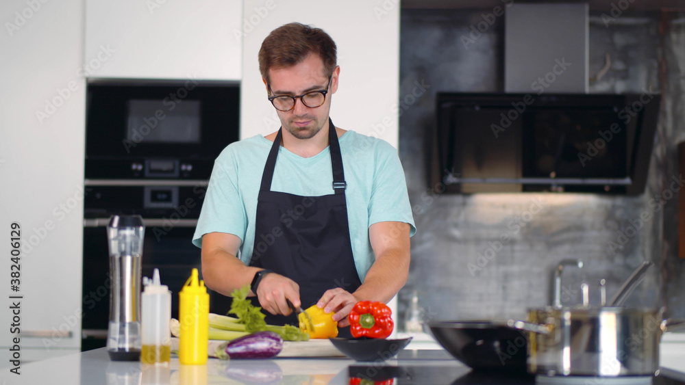 Cheerful young man in apron preparing healthy meal with vegetables in kitchen.
