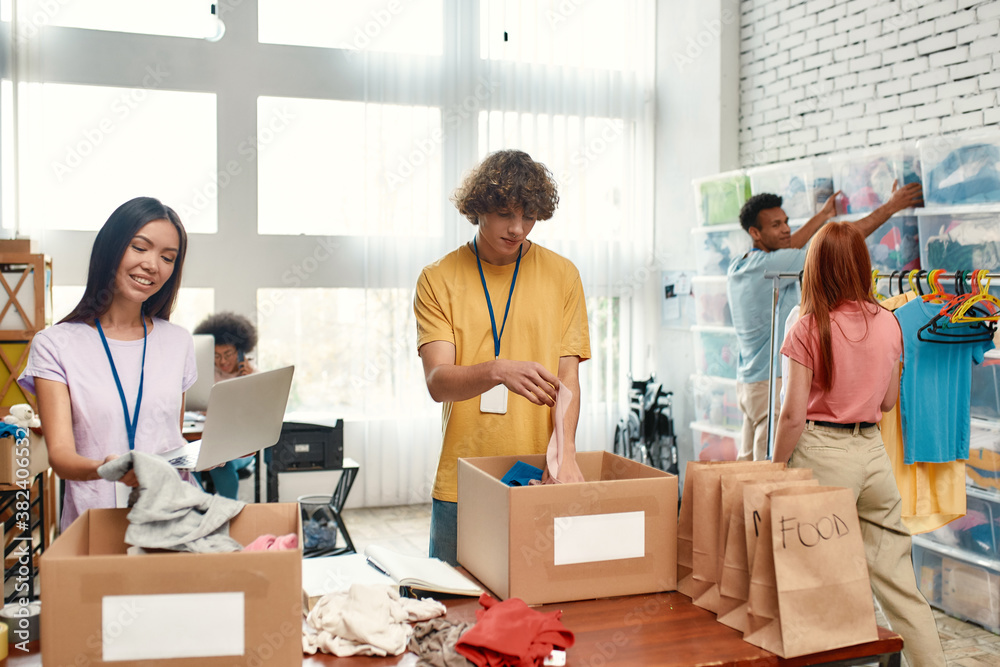 Fototapeta premium Young volunteers group sorting, packing food and clothes in cardboard boxes, Diverse team working on donation project indoors