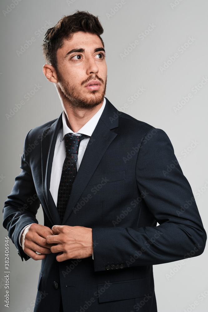 Young man with beard wearing a dark blue suit. He is fastening the suit jacket while looks to his left. Young man with beard wearing a dark blue suit. He is fastening the suit jacket while looks to his left.