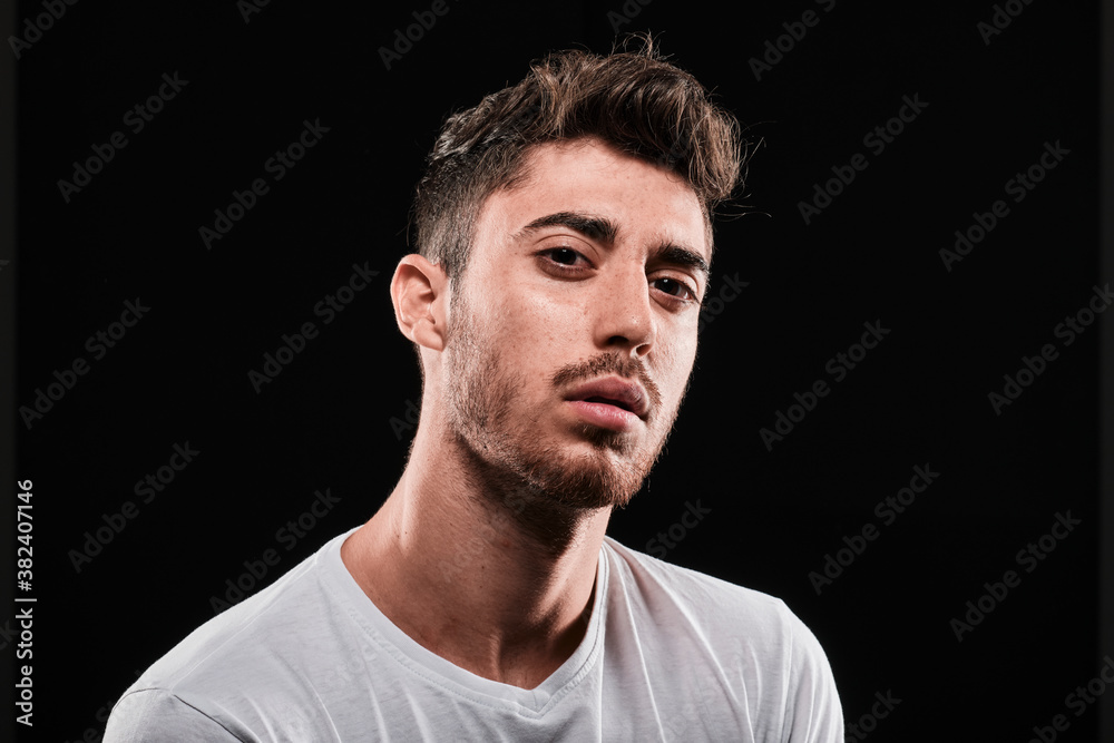 Young man close up portrait on a black background. He is wearing a basic white tshirt and looking at camera.