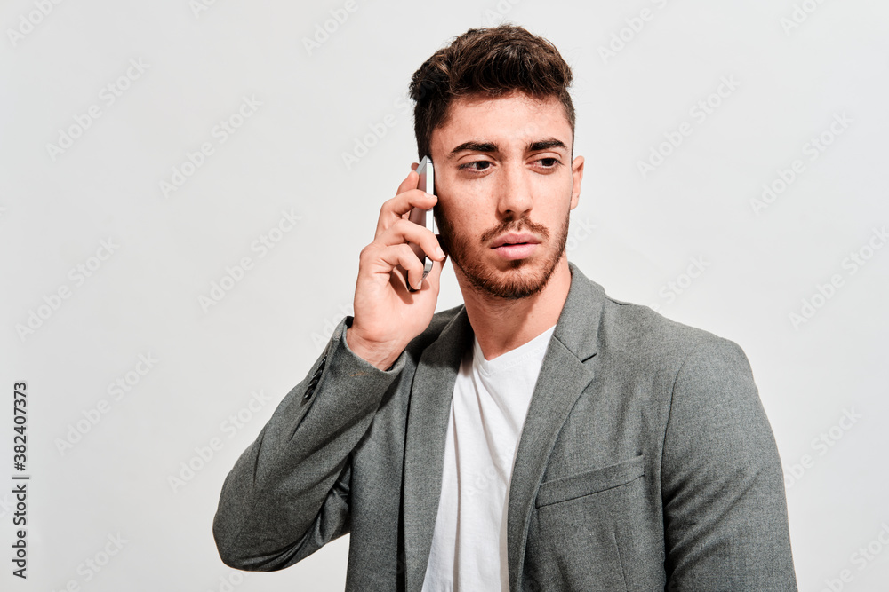 Serious young man wearing jeans, a white tshirt and a grey blazer walking in front of a white wall while talking with someone on his mobile phone Serious young man wearing jeans, a white tshirt and a grey blazer walking in front of a white wall while talking with someone on his mobile phone