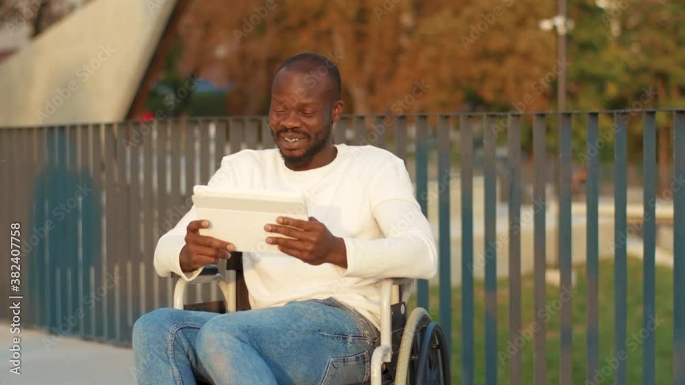 Smiling young african american man on a wheelchair with tablet computer in park at sunlight disability medicine. Possibility handicapped handicap internet. Slow motion Smiling young african american man on a wheelchair with tablet computer in park at sunlight disability medicine. Possibility handicapped handicap internet. Slow motion